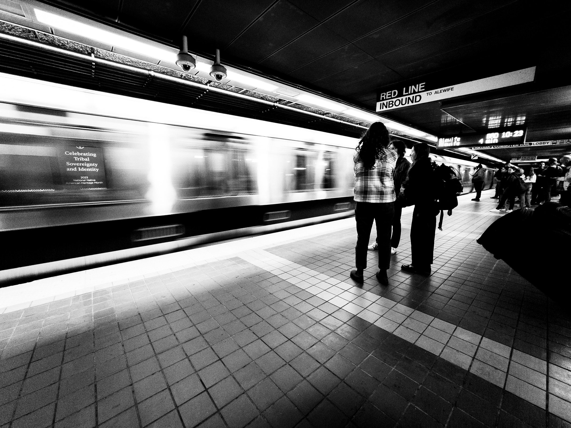 A black and white photograph of a subway station platform. A train is speeding past, creating a blur. Two people stand on the platform, facing the train. One person is wearing a plaid shirt and the other is wearing a dark jacket. Other people are standing further down the platform. A sign above reads "Red Line Inbound to Alewife." The platform is tiled and the ceiling is lined with lights.