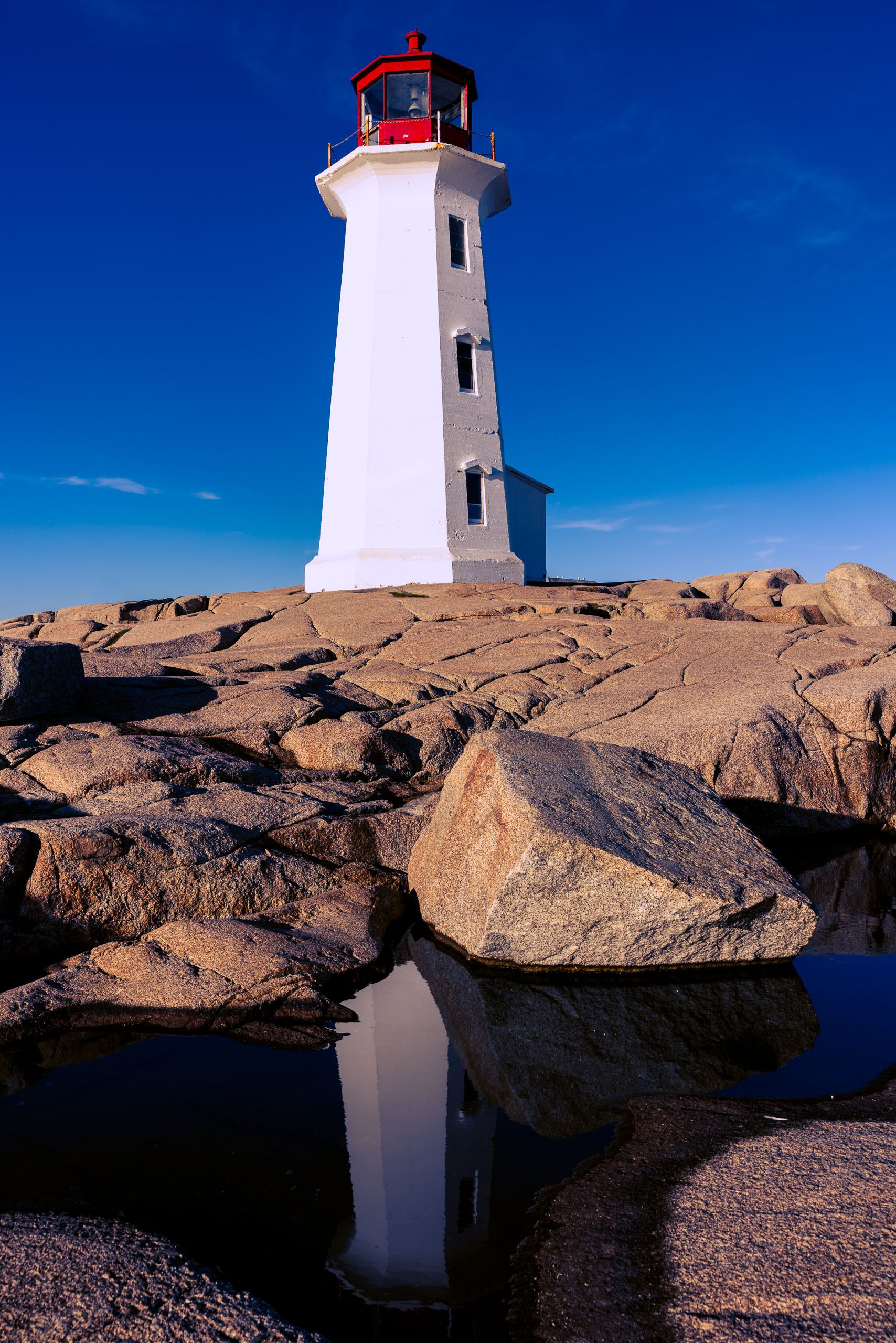 A tall, white lighthouse with a red top stands on rugged, light-colored rocks. The sky is clear and blue, with a few wispy clouds. In the foreground, a pool of water reflects the lighthouse, creating a mirror-like effect. The scene conveys a sense of isolation and tranquility, typical of coastal landscapes.