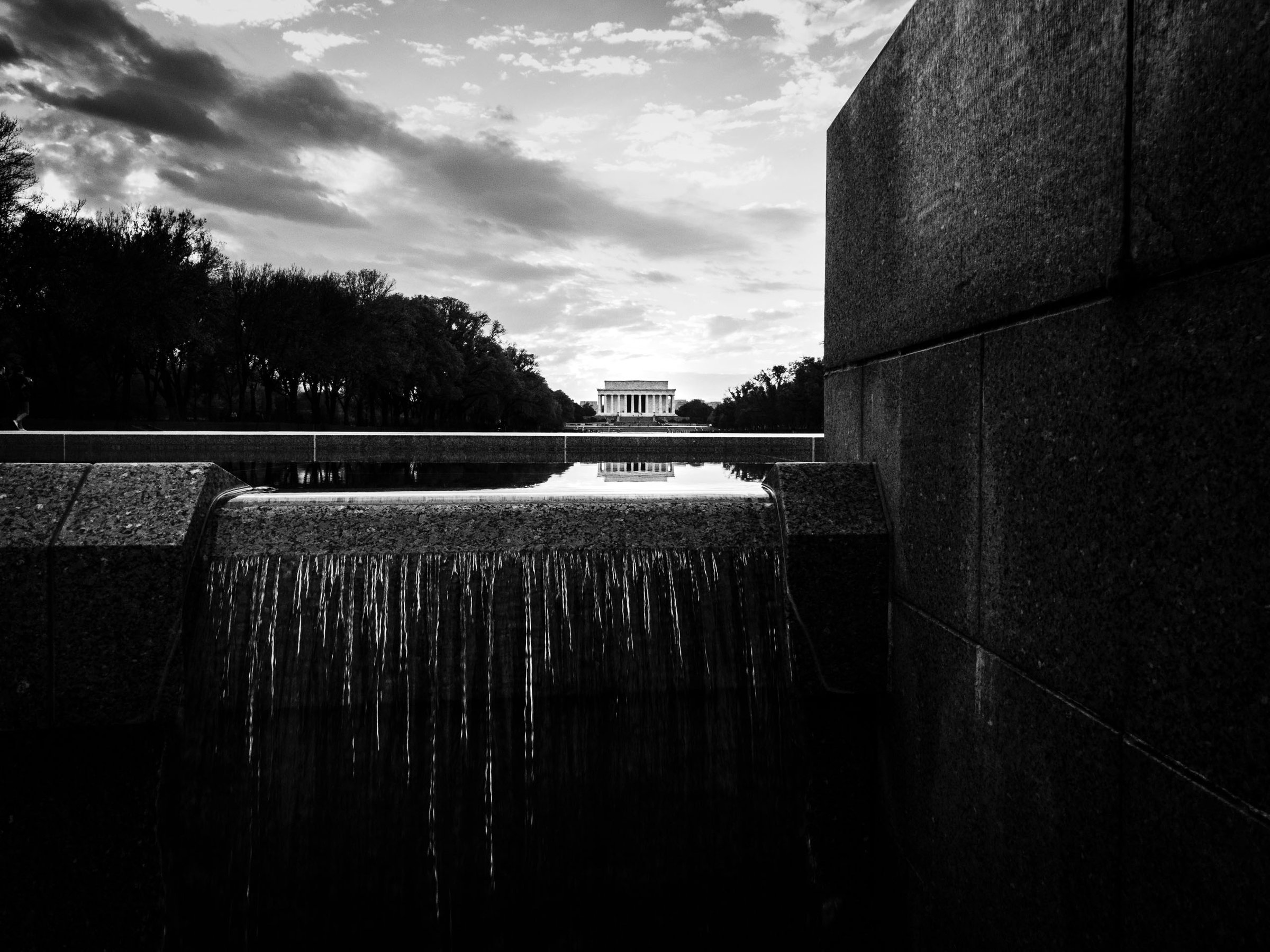 A black and white photograph shows a serene scene with the Lincoln Memorial in the distance and its reflection in the water of the pool of the World War II Memorial. In the foreground, water spills over an edge of the granite of the World War II Memorial. The sky is filled with scattered clouds, creating a dramatic backdrop. Tall, dark trees line both sides of the pool, adding to the symmetrical and tranquil atmosphere. The foreground features large, textured stone walls of the World War II Memorial, framing the view.