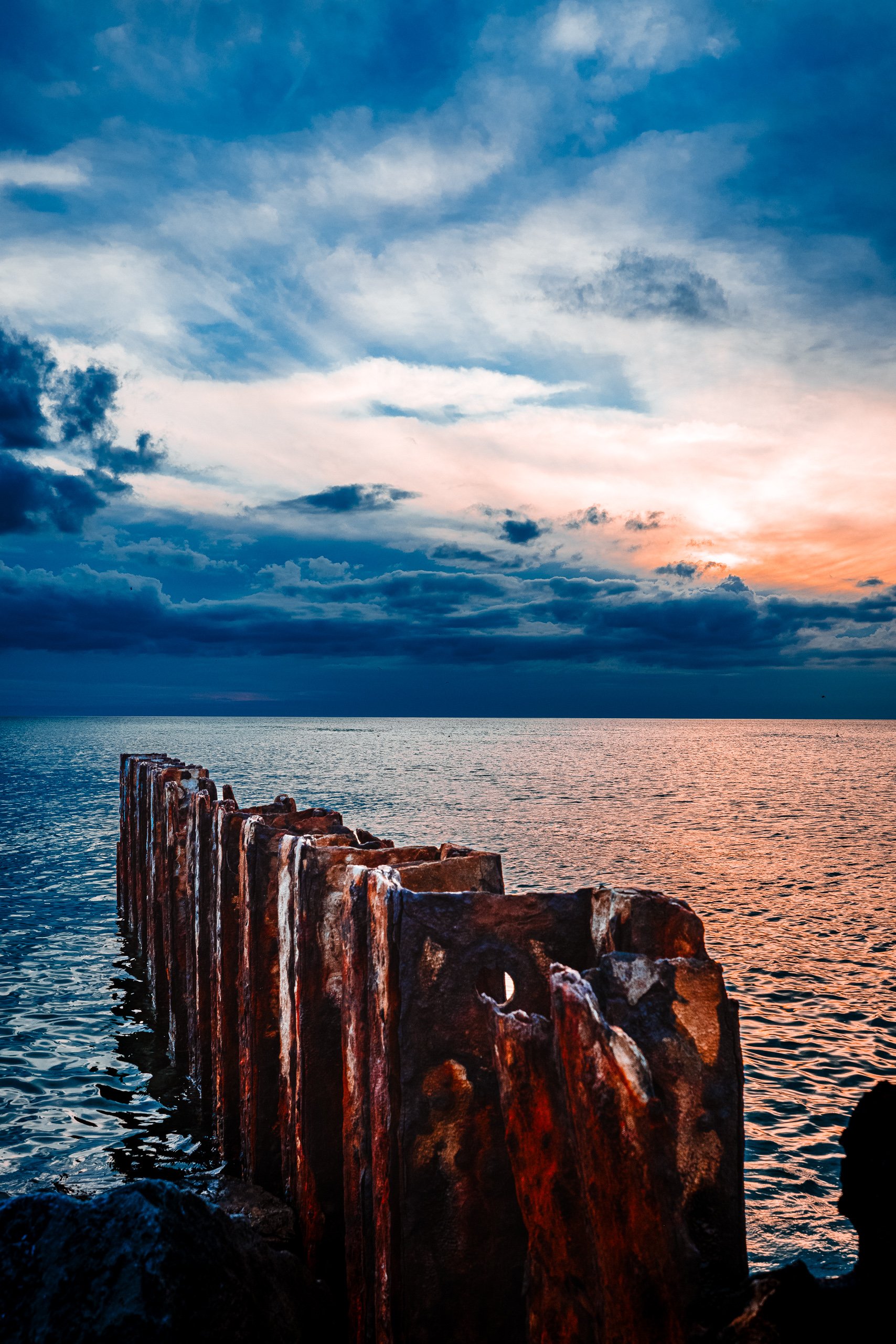 A rugged, weathered, and rusted sea wall extends into a calm sea under a dramatic sky. The sky is a blend of deep blues and warm oranges, in a sunset. Wispy clouds stretch across the horizon, catching the light. The water reflects the sky's colors, creating a serene and picturesque scene. The sea wall's rough, textured surface contrasts with the smooth water, adding a sense of age and resilience to the landscape.