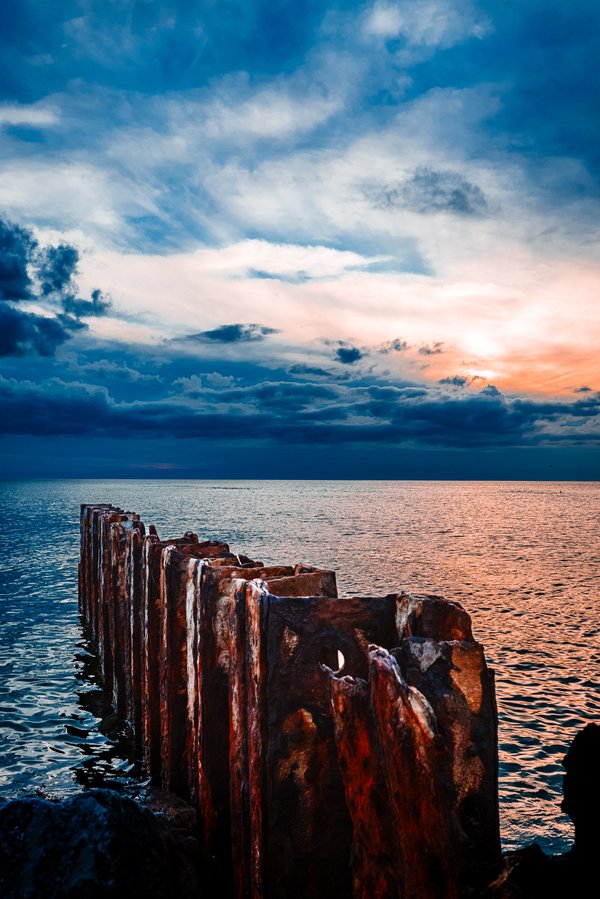 A rugged, weathered, and rusted sea wall extends into a calm sea under a dramatic sky. The sky is a blend of deep blues and warm oranges, in a sunset. Wispy clouds stretch across the horizon, catching the light. The water reflects the sky's colors, creating a serene and picturesque scene. The sea wall's rough, textured surface contrasts with the smooth water, adding a sense of age and resilience to the landscape.