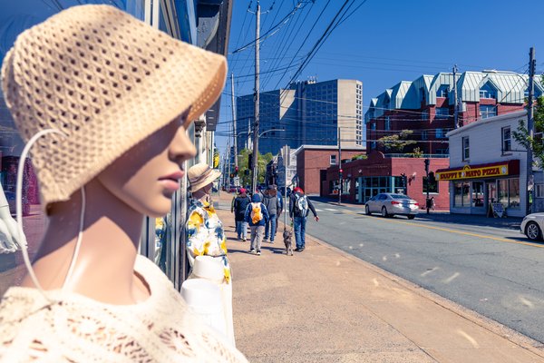 A mannequin wearing a straw hat and a white knitted top stands in a store window. Outside, people walk along a city street lined with brick and white buildings. A pizza shop with a yellow sign is visible. Cars drive on the road, and modern high-rises rise in the background under a clear blue sky. Power lines crisscross above, and a few trees add greenery to the urban scene.