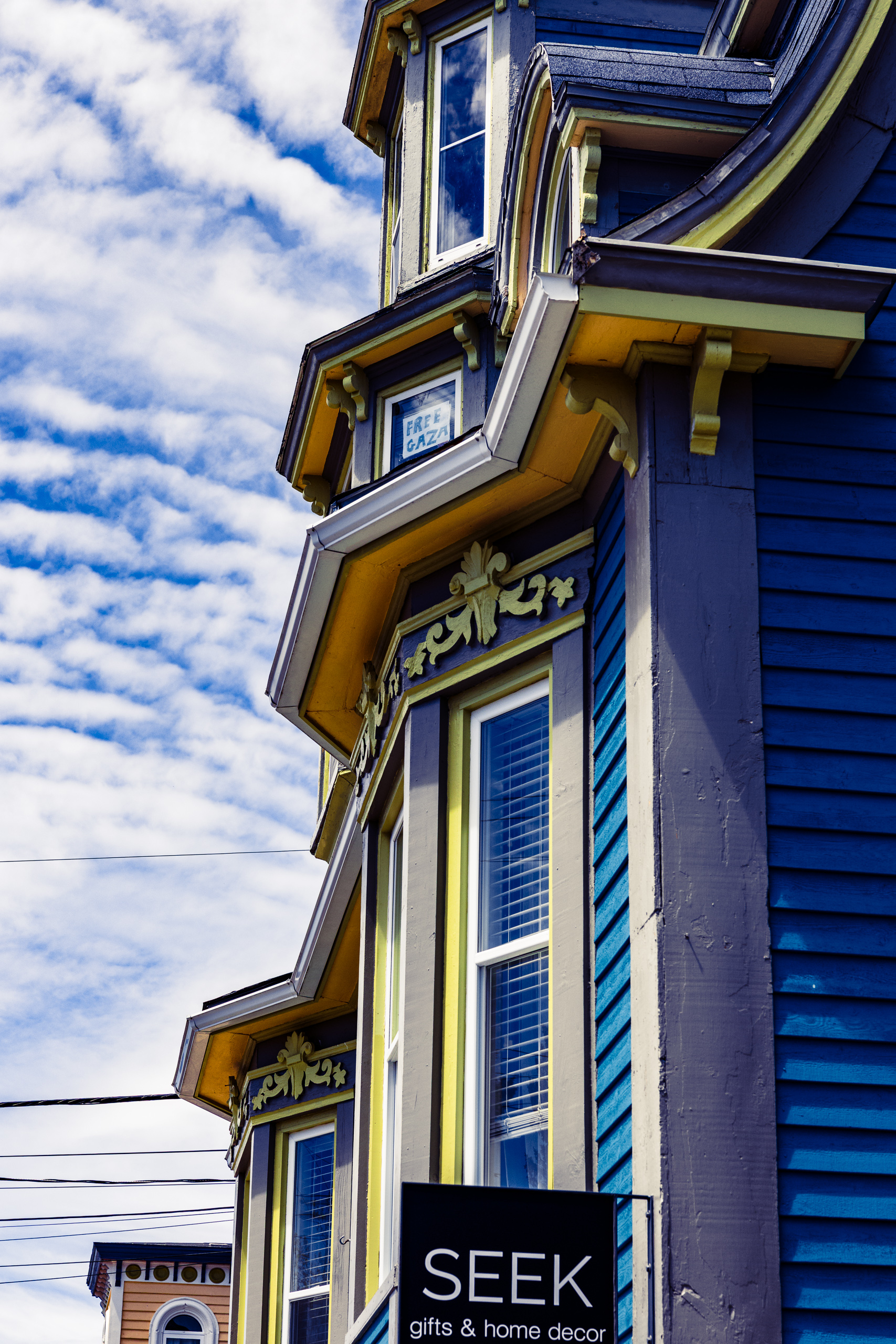 A vibrant Victorian-style building features a striking color palette of deep blue, yellow, and purple. The facade includes ornate trim and bay windows, adding architectural interest. A sign in front reads "SEEK gifts & home decor," indicating a retail space. A sign in an upper floor window reads 'FREE GAZA'. The sky is bright with scattered white clouds, enhancing the vivid colors of the building. Power lines are visible in the foreground, suggesting an urban setting.