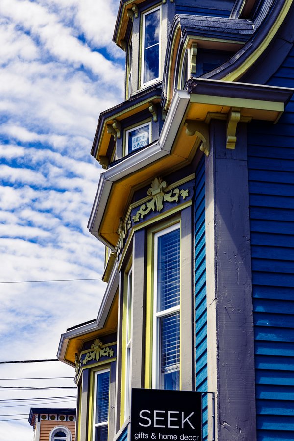 A vibrant Victorian-style building features a striking color palette of deep blue, yellow, and purple. The facade includes ornate trim and bay windows, adding architectural interest. A sign in front reads "SEEK gifts & home decor," indicating a retail space. A sign in an upper floor window reads 'FREE GAZA'. The sky is bright with scattered white clouds, enhancing the vivid colors of the building. Power lines are visible in the foreground, suggesting an urban setting.