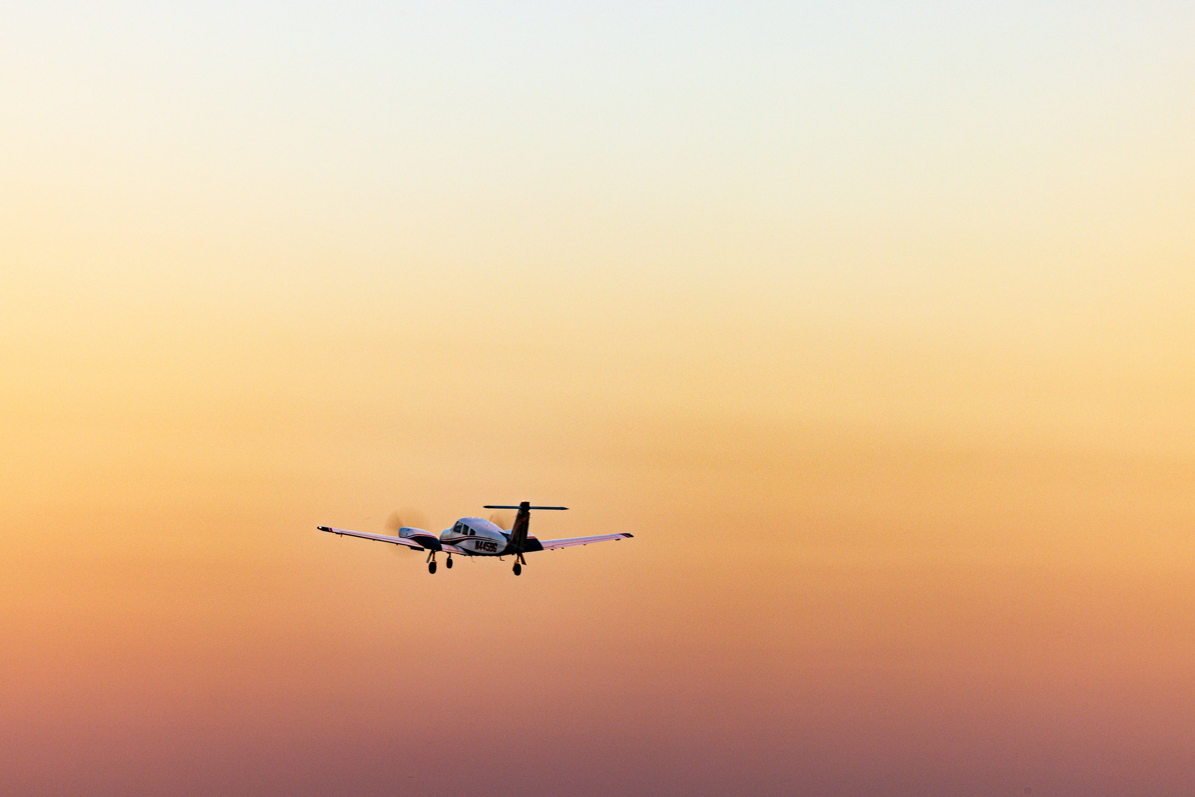 A small propeller airplane flies against a gradient sky transitioning from pale yellow at the top to deep orange near the horizon. The aircraft is silhouetted, with its landing gear extended, suggesting it is either approaching or departing. The scene conveys a sense of calm and tranquility, typical of early morning or late afternoon lighting. The airplane's position slightly off-center adds dynamic balance to the composition.
