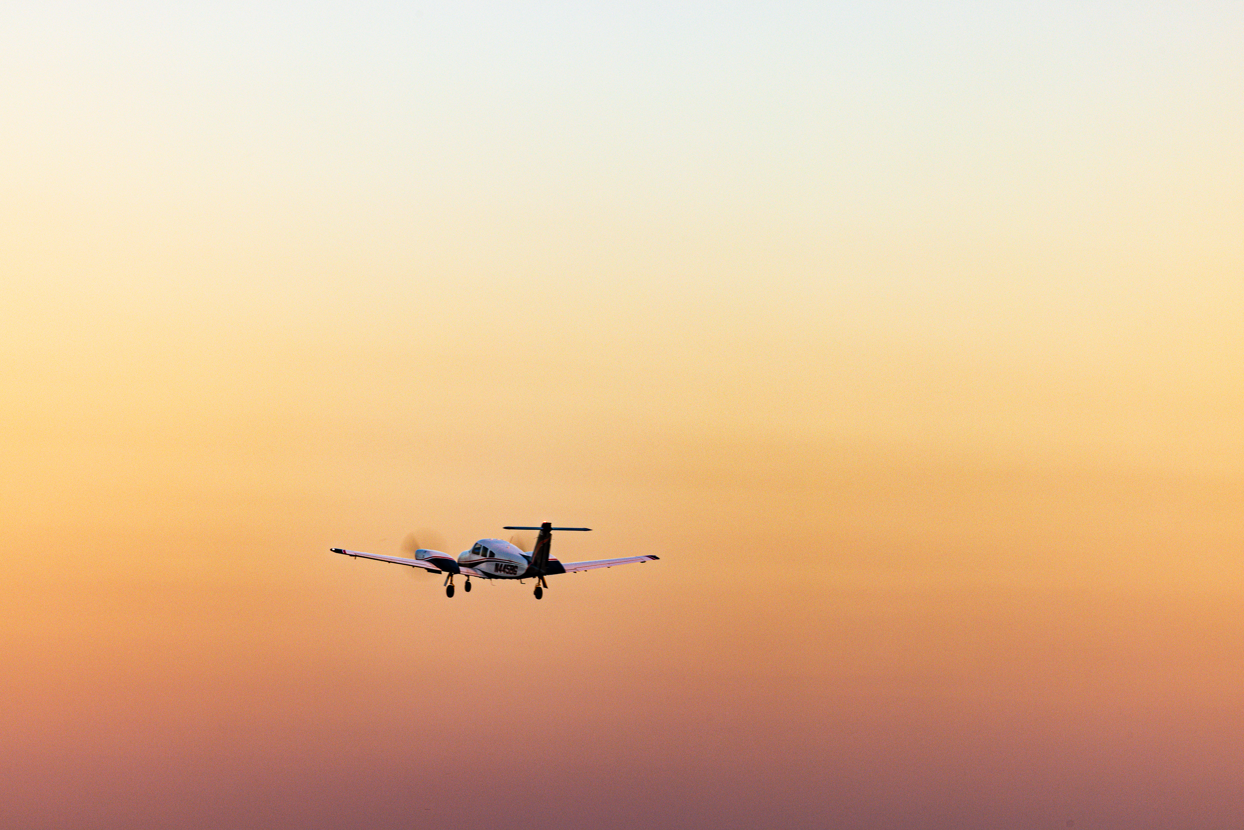 A small propeller airplane flies against a gradient sky transitioning from pale yellow at the top to deep orange near the horizon. The aircraft is silhouetted, with its landing gear extended, suggesting it is either approaching or departing. The scene conveys a sense of calm and tranquility, typical of early morning or late afternoon lighting. The airplane's position slightly off-center adds dynamic balance to the composition.