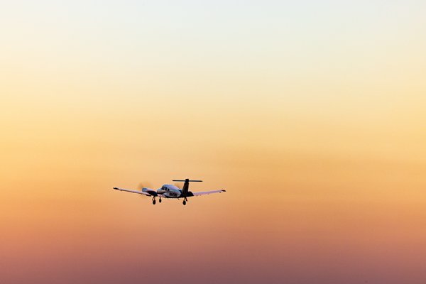 A small propeller airplane flies against a gradient sky transitioning from pale yellow at the top to deep orange near the horizon. The aircraft is silhouetted, with its landing gear extended, suggesting it is either approaching or departing. The scene conveys a sense of calm and tranquility, typical of early morning or late afternoon lighting. The airplane's position slightly off-center adds dynamic balance to the composition.