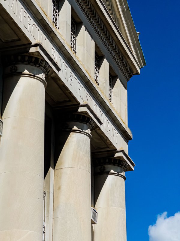 Low-angle upward view of the limestone Beaux-Arts facade of the Commercial Bank & Trust Company building, in Bowling Green, Ohio, showing massive fluted columns, dentil cornice molding, leaded geometric transom windows, and partially visible incised lettering against a deep blue September sky with a small cloud at lower right.