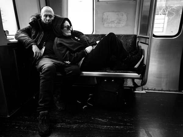 A black and white photo shows two people sitting on a subway seat. The man on the left, wearing a fur-lined jacket, leans back with an arm around the other. The woman on the right, in a hooded jacket and sunglasses, rests their head on the man's shoulder, appearing to sleep. The subway car has graffiti on the walls and windows, and the floor is dark and worn. The scene conveys a sense of urban life and quiet companionship.