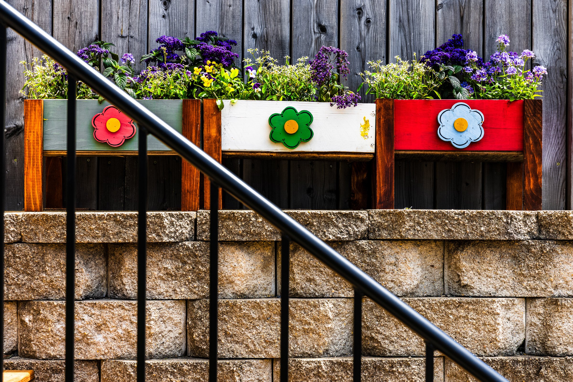 Three wooden planters with vibrant flowers sit on a stone ledge. Each planter is painted a different color: green, white, and red. They are adorned with large, colorful flower decorations in matching hues—red, green, and blue. Behind the planters, a dark wooden fence provides a rustic backdrop. A black metal railing runs diagonally across the foreground, adding structure to the scene. The flowers are lush and varied, featuring shades of purple, yellow, and green, creating a lively and cheerful display.