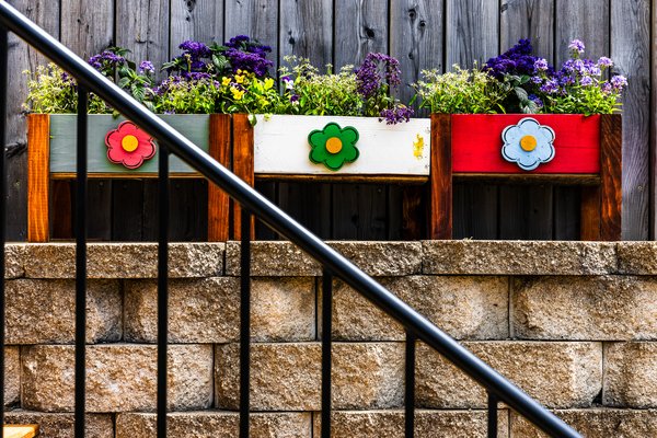 Three wooden planters with vibrant flowers sit on a stone ledge. Each planter is painted a different color: green, white, and red. They are adorned with large, colorful flower decorations in matching hues—red, green, and blue. Behind the planters, a dark wooden fence provides a rustic backdrop. A black metal railing runs diagonally across the foreground, adding structure to the scene. The flowers are lush and varied, featuring shades of purple, yellow, and green, creating a lively and cheerful display.
