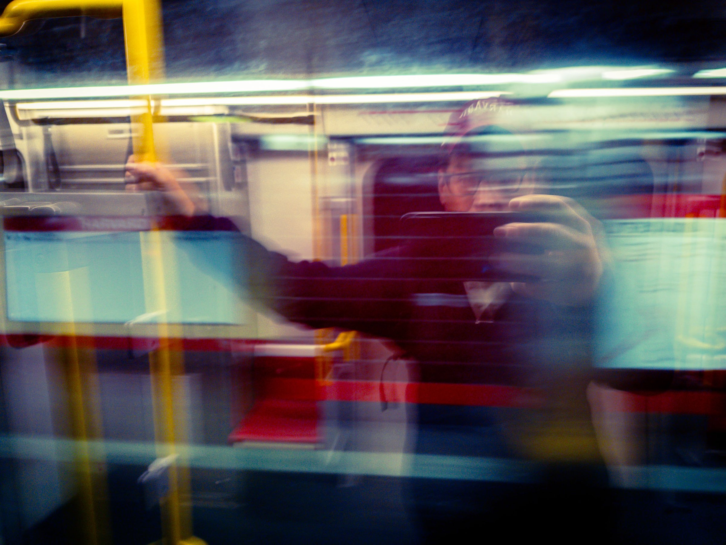A blurred scene inside a moving train. A person in a dark jacket stands, holding a smartphone, with motion lines indicating movement. The interior features red and gray seats, and overhead lighting casts a soft glow. Other passengers are partially visible, also blurred, suggesting a busy commute. The overall atmosphere conveys the dynamic and transient nature of public transportation.