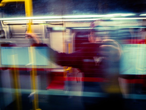 A blurred scene inside a moving train. A person in a dark jacket stands, holding a smartphone, with motion lines indicating movement. The interior features red and gray seats, and overhead lighting casts a soft glow. Other passengers are partially visible, also blurred, suggesting a busy commute. The overall atmosphere conveys the dynamic and transient nature of public transportation.