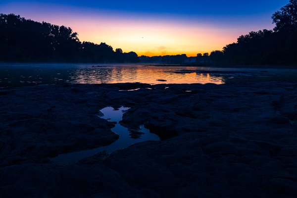 A river at dawn glows orange and purple between dark tree-lined banks, with mist rising over the water and exposed rock ledges in the foreground.