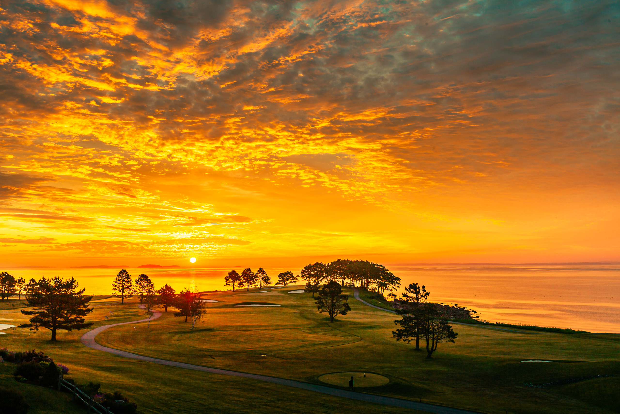 A vibrant sunrise paints the sky with fiery oranges and deep blues, casting a warm glow over a serene landscape. Silhouetted trees dot the rolling hills of a golf course, with winding paths leading to distant greens. The sun dips below the horizon, reflecting its golden light across a calm body of water. A wooden fence in the foreground adds rustic charm, enhancing the tranquil and picturesque scene.