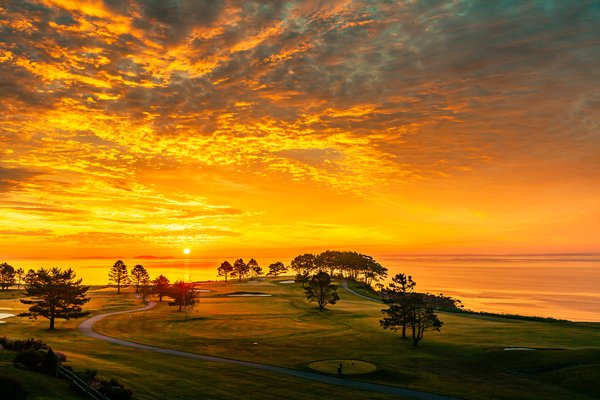 A vibrant sunrise paints the sky with fiery oranges and deep blues, casting a warm glow over a serene landscape. Silhouetted trees dot the rolling hills of a golf course, with winding paths leading to distant greens. The sun dips below the horizon, reflecting its golden light across a calm body of water. A wooden fence in the foreground adds rustic charm, enhancing the tranquil and picturesque scene.