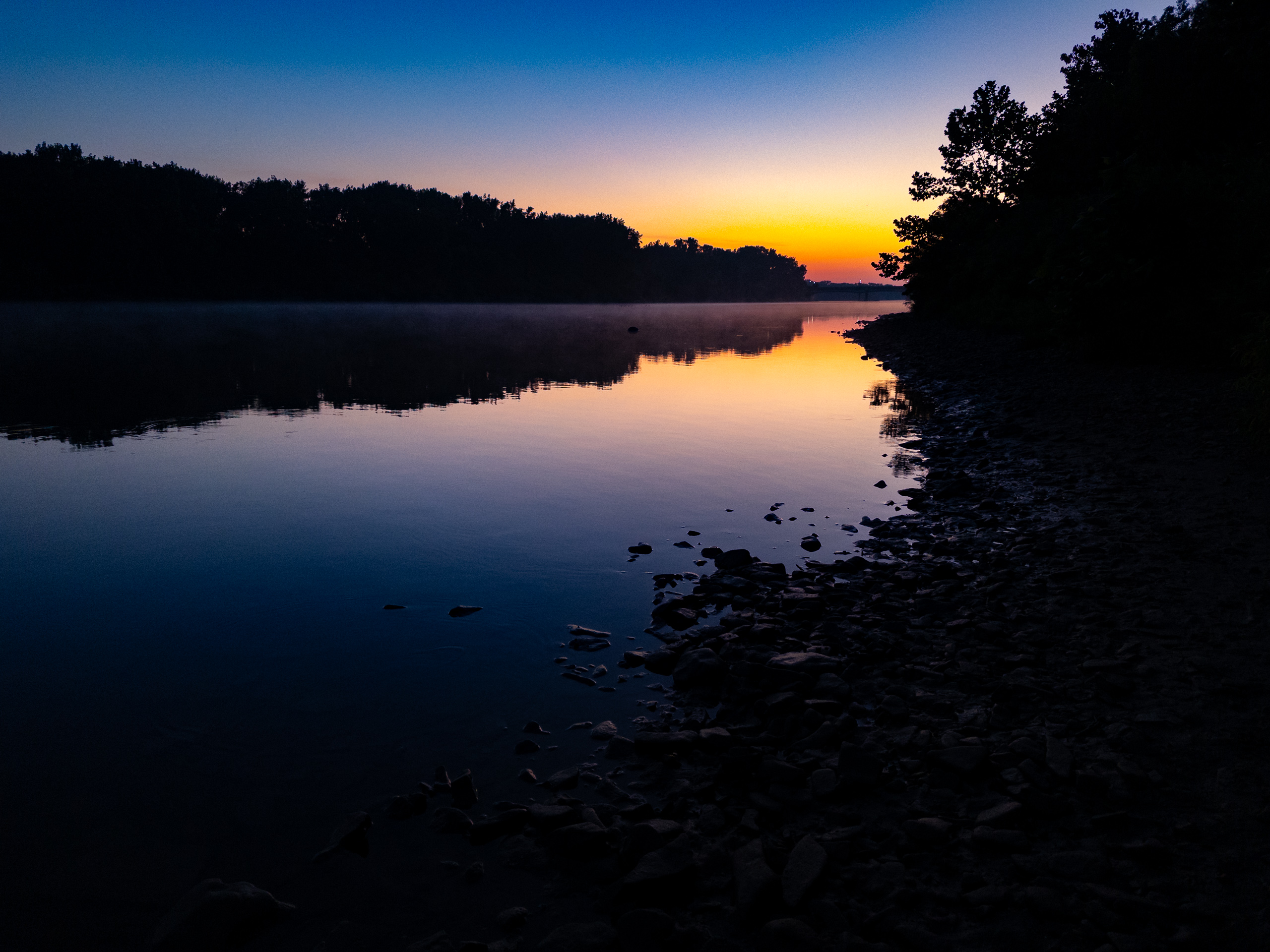 Pre-dawn twilight over a calm river, with a glowing amber and orange horizon fading upward into deep blue sky. Tree silhouettes line both banks. Light morning mist drifts above the glassy water, which mirrors the sky's gradient. A rocky shoreline extends along the right foreground into shadow.