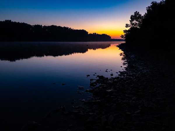Pre-dawn twilight over a calm river, with a glowing amber and orange horizon fading upward into deep blue sky. Tree silhouettes line both banks. Light morning mist drifts above the glassy water, which mirrors the sky's gradient. A rocky shoreline extends along the right foreground into shadow.