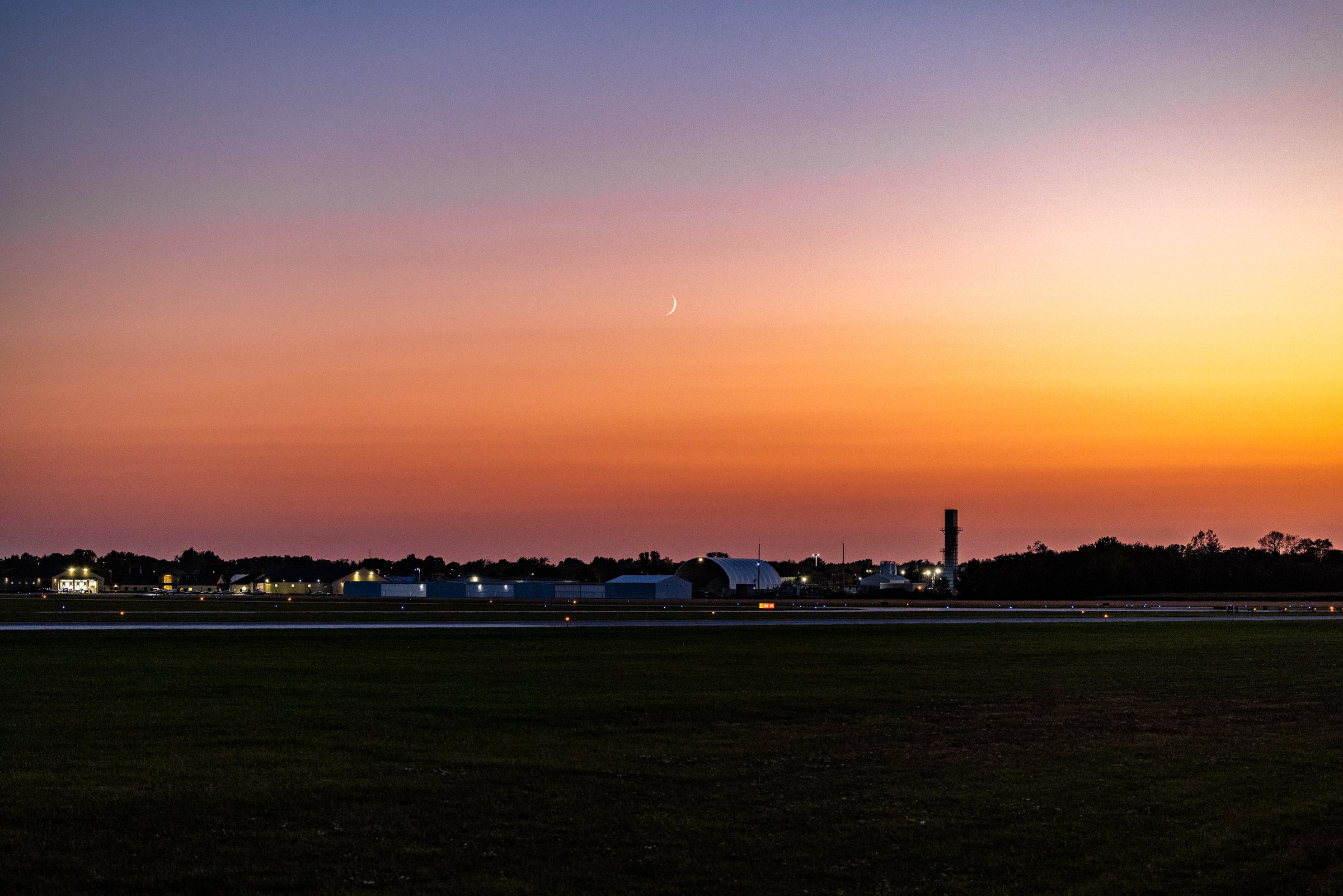 A serene sunset over an airport runway. The sky transitions from deep blue at the top to vibrant orange and pink near the horizon. Silhouetted against the colorful sky are several hangars and airport buildings, with a few lights glowing softly. A crescent moon and a bright star are visible in the sky. The foreground features a grassy field adjacent to the runway, creating a peaceful and expansive scene.
