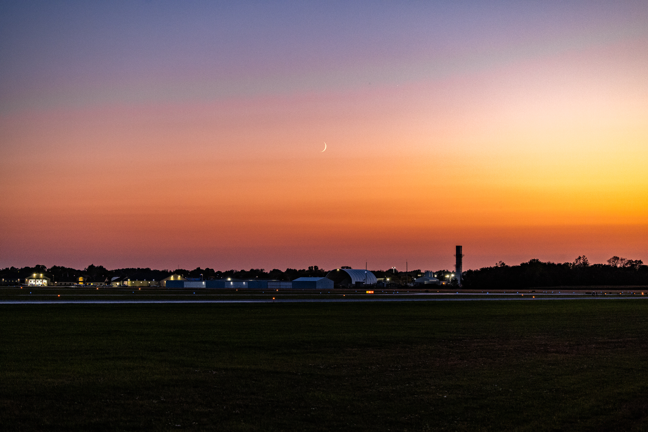 A serene sunset over an airport runway. The sky transitions from deep blue at the top to vibrant orange and pink near the horizon. Silhouetted against the colorful sky are several hangars and airport buildings, with a few lights glowing softly. A crescent moon and a bright star are visible in the sky. The foreground features a grassy field adjacent to the runway, creating a peaceful and expansive scene.