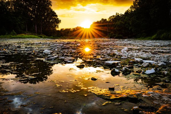 A brilliant sunburst sets directly on the horizon between two tree lines above a shallow, rocky river bed, its starburst rays and golden sky reflected in a still pool of water in the foreground among moss-covered stones in the river.