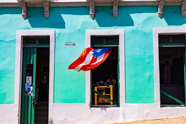 A Puerto Rican flag billows outward from an open window in a turquoise-painted colonial building at 500 Norzagaray Street in Old San Juan, Puerto Rico. A figure is visible inside the darkened interior.