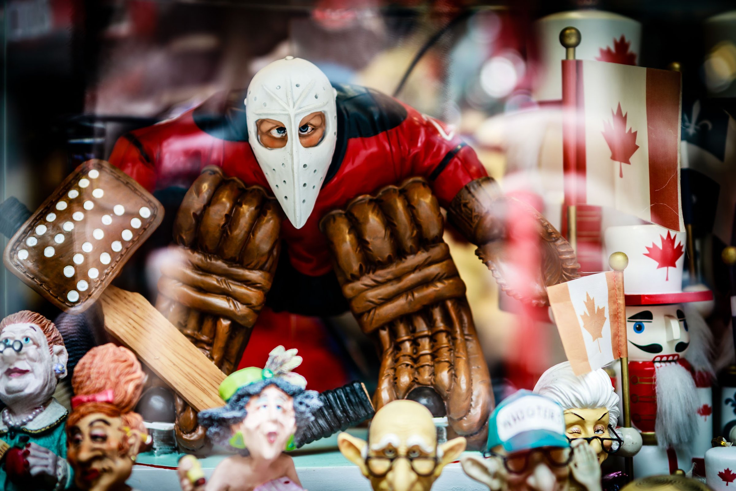 A collection of colorful figurines in a display case. The central figure is an ice hockey goalie with a white face mask. Surrounding the goalie are various characters, including royalty, a toy soldier, and a Canadian flag. The figurines are detailed and vibrant, with expressive faces and intricate costumes. The display is lively and colorful showcasing an overall theme of Canadian pride.