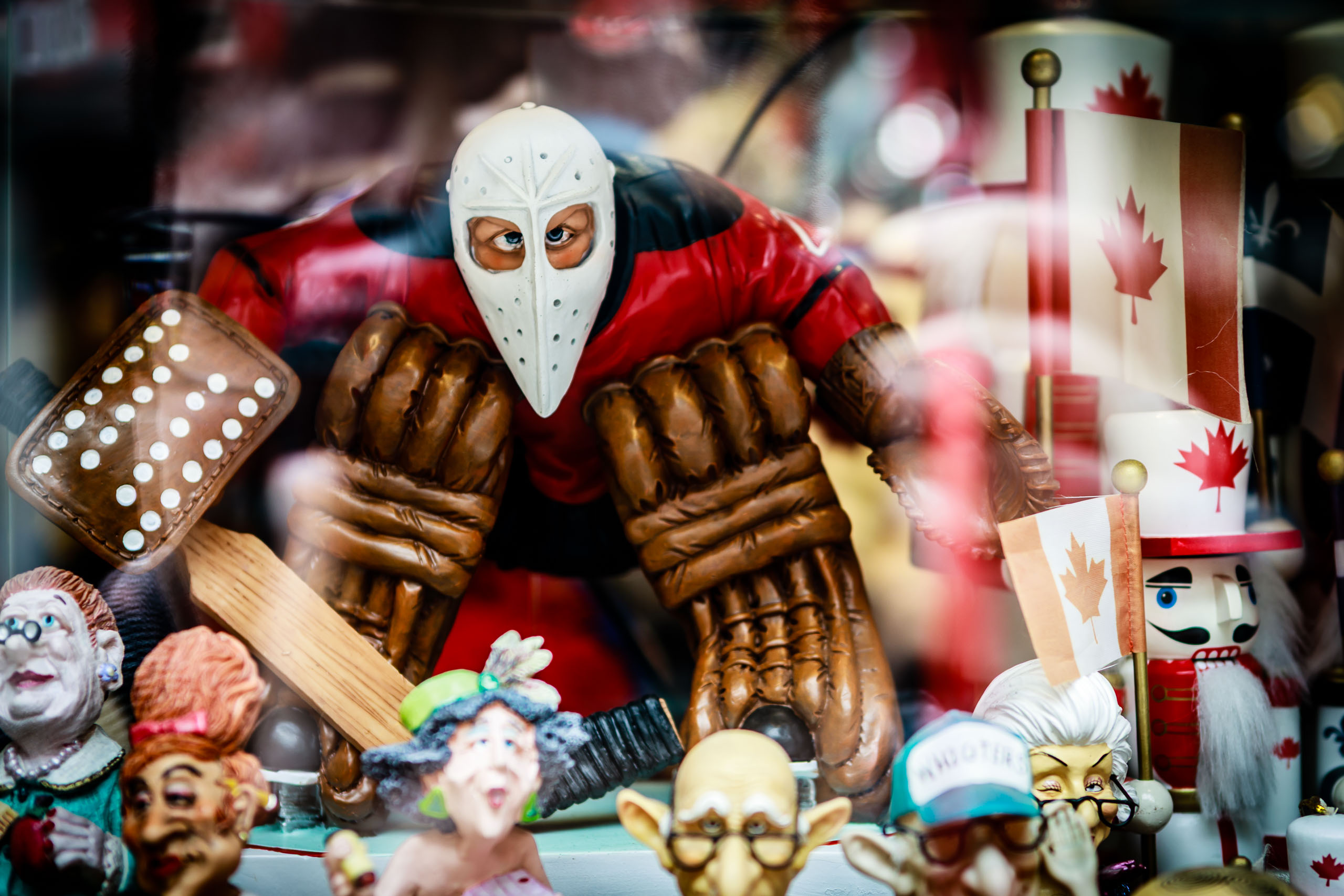 A collection of colorful figurines in a display case. The central figure is an ice hockey goalie with a white face mask. Surrounding the goalie are various characters, including royalty, a toy soldier, and a Canadian flag. The figurines are detailed and vibrant, with expressive faces and intricate costumes. The display is lively and colorful showcasing an overall theme of Canadian pride.