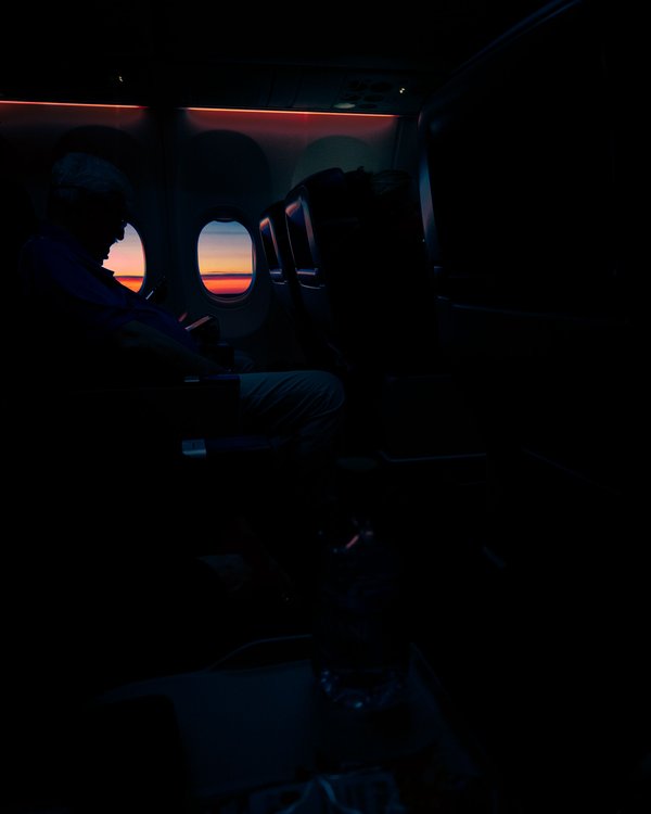 A person sits in an airplane seat, silhouetted against a vibrant sunset visible through the window. The sky transitions from deep orange to soft pink, casting a warm glow inside the cabin. The passenger is focused on the screen of a mobile phone. The interior is dimly lit, with overhead compartments and seat details faintly visible. The scene conveys a sense of calm and travel during twilight.