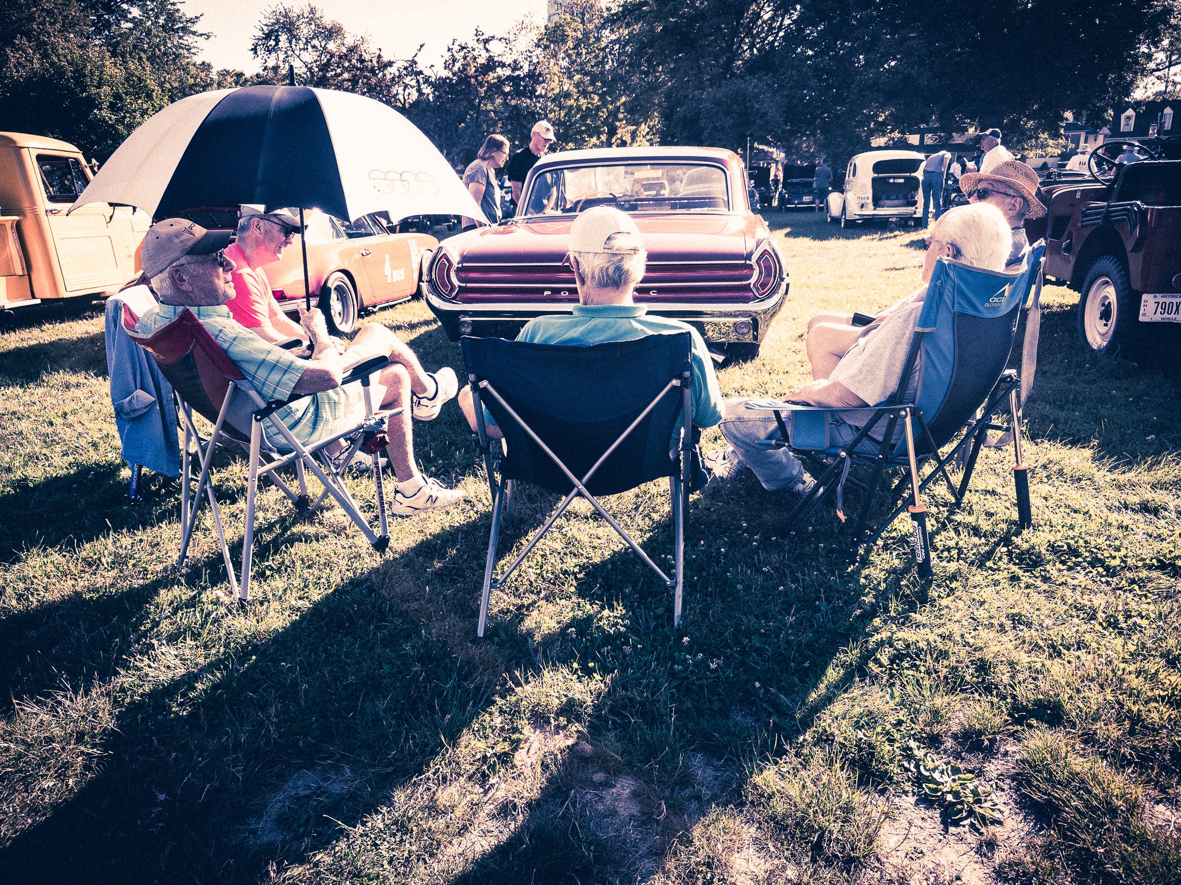 A group of elderly men relax in folding chairs under a large umbrella on a grassy field. They are facing vintage cars, including a prominent orange one with chrome details. The scene is sunny, with shadows cast on the ground. Trees and more classic vehicles are visible in the background. The people appear to be enjoying a leisurely day at a car show or outdoor gathering, dressed in casual summer clothing and hats.