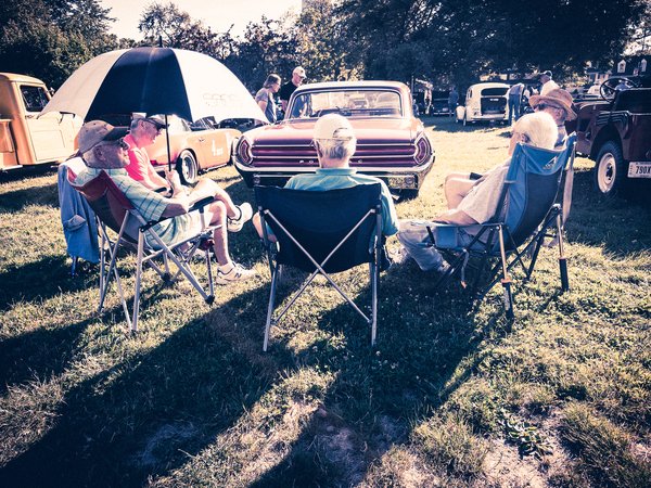 A group of elderly men relax in folding chairs under a large umbrella on a grassy field. They are facing vintage cars, including a prominent orange one with chrome details. The scene is sunny, with shadows cast on the ground. Trees and more classic vehicles are visible in the background. The people appear to be enjoying a leisurely day at a car show or outdoor gathering, dressed in casual summer clothing and hats.