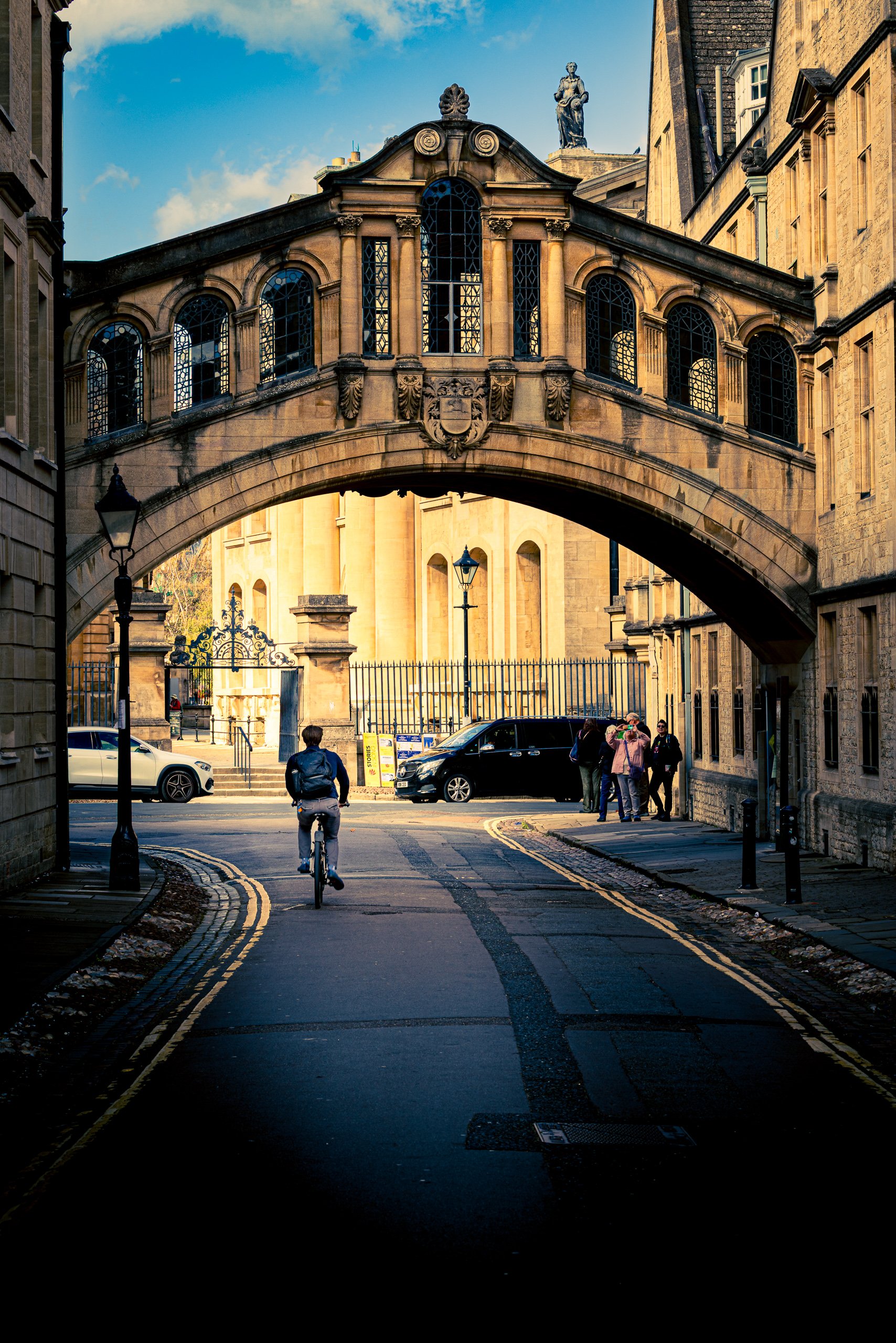 A stone bridge arches over a cobblestone path, connecting historic buildings. The bridge features ornate windows and decorative stonework. Below, a cyclist rides towards the camera, flanked by pedestrians walking on either side. Cars are parked nearby, and a wrought iron gate leads to a grand entrance with statues. The scene is bathed in warm sunlight, casting long shadows and highlighting the architectural details. The sky is a clear blue with scattered clouds, adding to the picturesque setting.