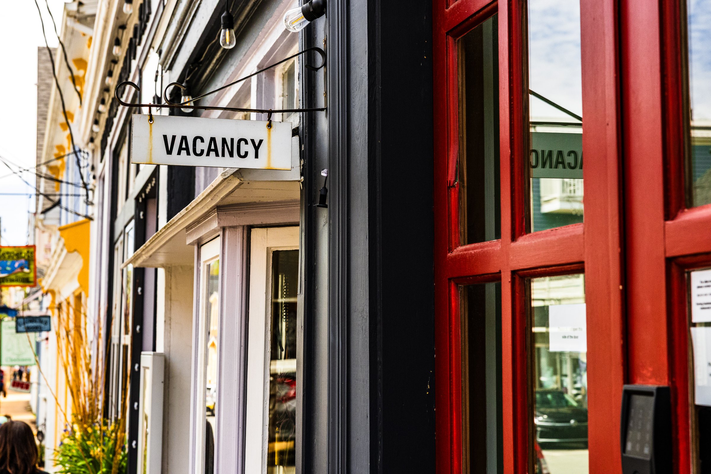 A storefront with a black and white "VACANCY" sign hanging above a glass door. The door is framed in bright red, contrasting with the dark exterior. Above, string lights are visible, adding a cozy ambiance. To the left, a glimpse of a yellow building and other storefronts can be seen along a bustling street. A potted plant with green and yellow foliage sits near the entrance, adding a touch of nature. The overall scene suggests a lively urban area with potential opportunities for new businesses or tenants.