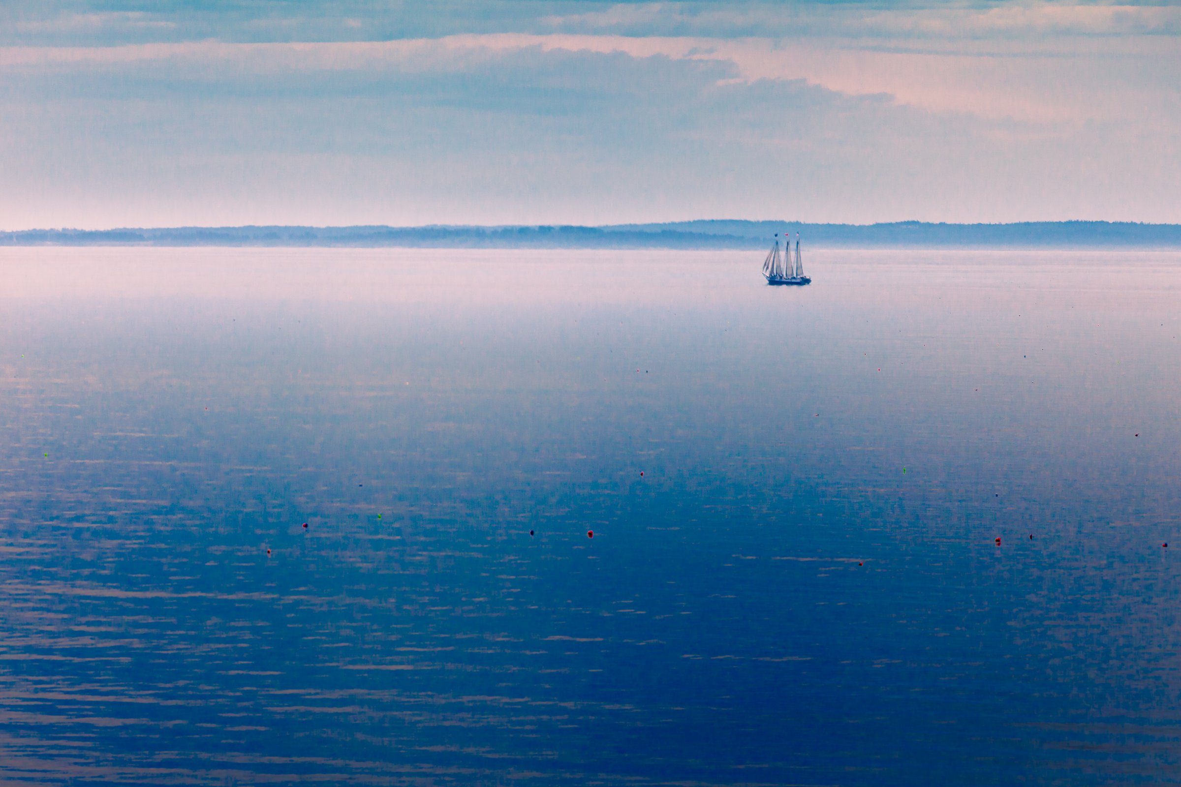 A serene seascape at dusk features a calm, expansive body of water reflecting soft pastel hues from the sky. A solitary sailboat with two masts glides gently across the horizon, silhouetted against the distant shoreline. The sky is dotted with wispy clouds, transitioning from light blue to hints of pink and purple. Scattered buoys float on the water's surface, adding subtle color accents. The overall atmosphere is tranquil and peaceful, evoking a sense of quiet solitude and natural beauty.