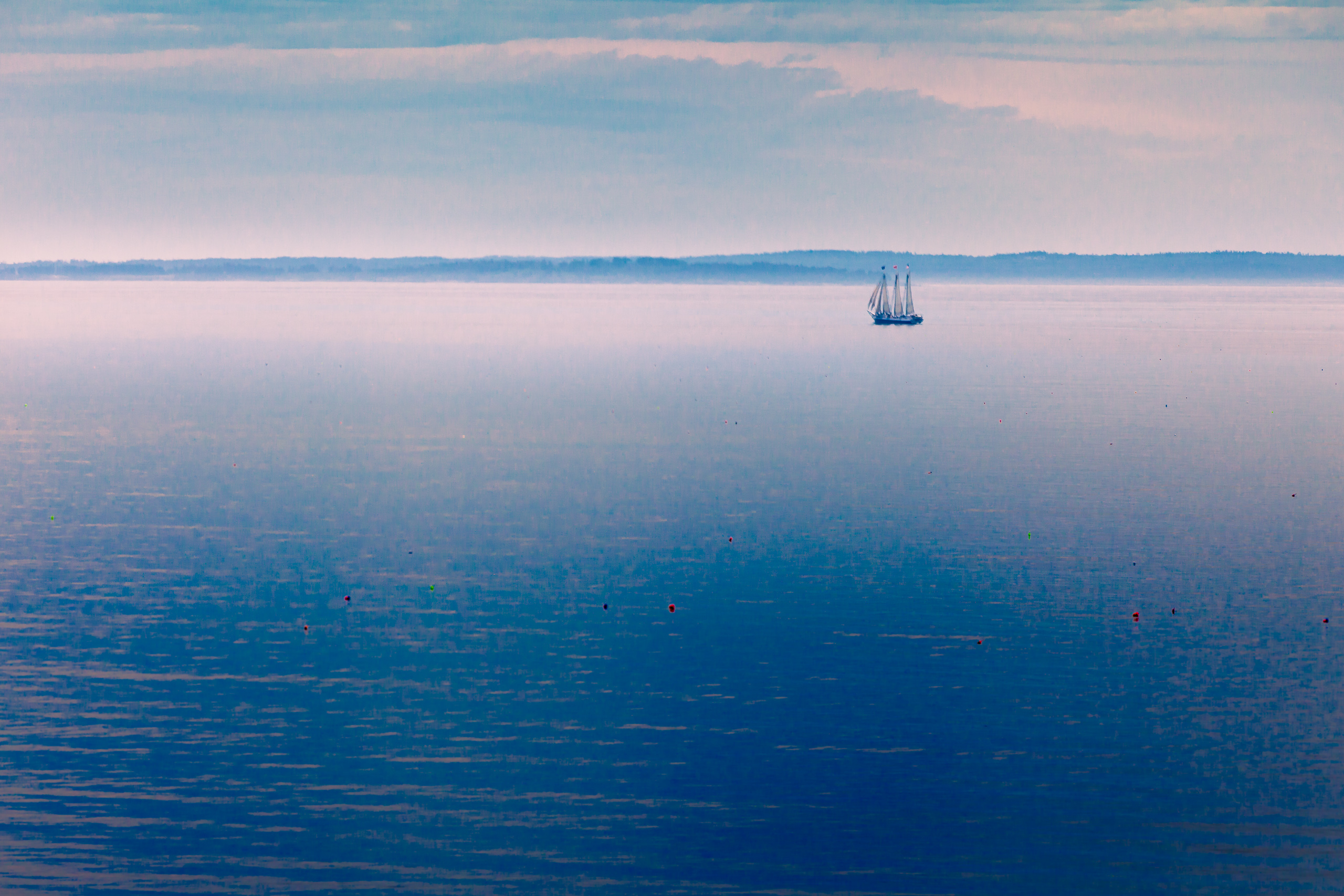 A serene seascape at dusk features a calm, expansive body of water reflecting soft pastel hues from the sky. A solitary sailboat with two masts glides gently across the horizon, silhouetted against the distant shoreline. The sky is dotted with wispy clouds, transitioning from light blue to hints of pink and purple. Scattered buoys float on the water's surface, adding subtle color accents. The overall atmosphere is tranquil and peaceful, evoking a sense of quiet solitude and natural beauty.