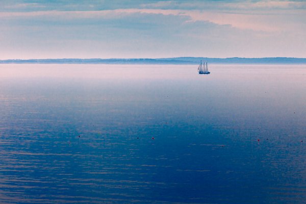 A serene seascape at dusk features a calm, expansive body of water reflecting soft pastel hues from the sky. A solitary sailboat with two masts glides gently across the horizon, silhouetted against the distant shoreline. The sky is dotted with wispy clouds, transitioning from light blue to hints of pink and purple. Scattered buoys float on the water's surface, adding subtle color accents. The overall atmosphere is tranquil and peaceful, evoking a sense of quiet solitude and natural beauty.