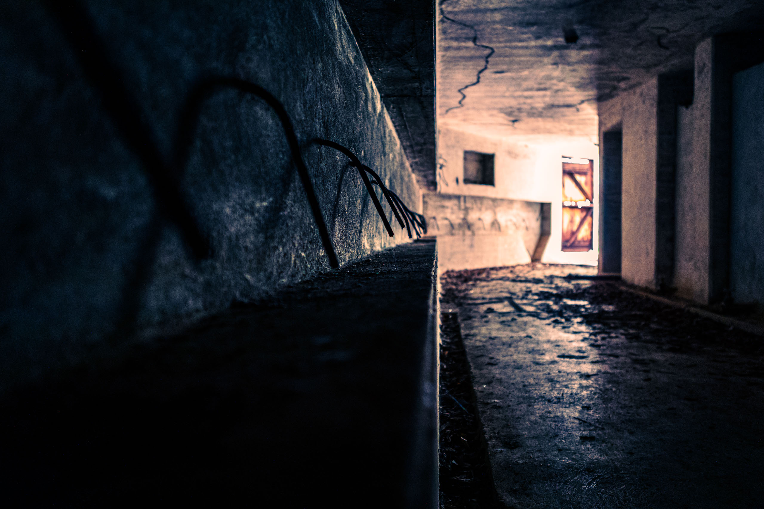 A dimly lit, concrete tunnel stretches into the distance. Shadows cast by a concrete ledge create abstract patterns on the walls. The floor appears to be damp, reflecting faint light. At the far end, a bright, warm glow emanates from an open door, contrasting with the cool, dark surroundings. The tunnel's walls are rough and cracked, adding to its abandoned, eerie atmosphere. The overall mood is mysterious and slightly unsettling, evoking a sense of exploration and the unknown.