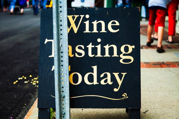 A black sign with gold lettering reads "Wine Tasting Today" on a metal post. The sign is positioned on a sidewalk next to a street. In the background, blurred figures of people walk by, some wearing red pants. The scene suggests a lively outdoor setting, possibly near a winery or event venue.